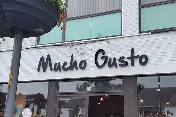 Witte bakstenen winkelgevel met zwarte sierlijke gevelletters "Mucho Gusto" boven de glazen entree van een horecapand.