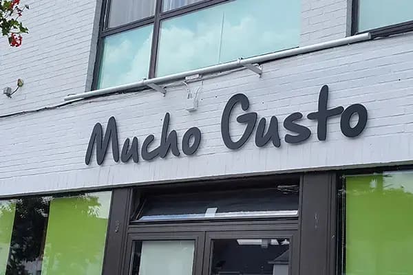 Witte bakstenen gevel met zwarte sierlijke gevelletters "Mucho Gusto" boven een glazen entree, toegepast als opvallende gevelreclame op een winkelpand.