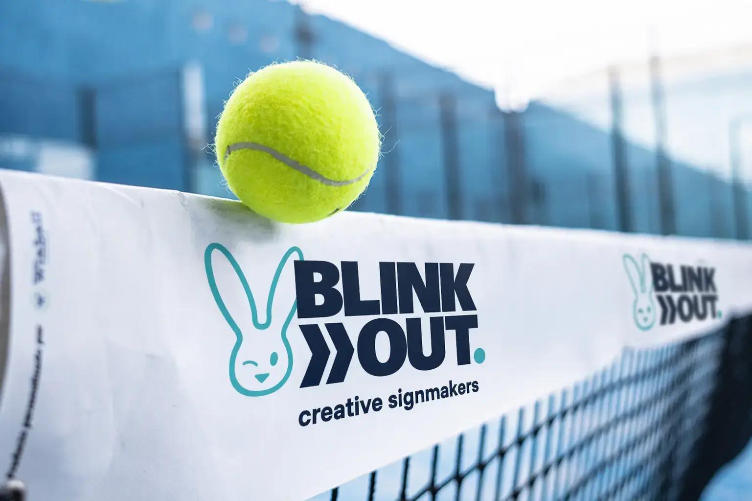 Witte netband met Blink Out creative signmakers-logo en turquoise beeldmerk op een tennisnet, met tennisbal op de rand en onscherpe sportbaan op de achtergrond.