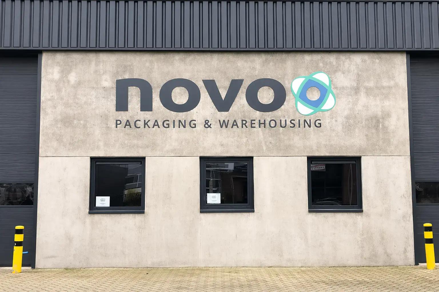 Bedrijfsgevel met groot novo-logo en bedrijfsbelettering Packaging & Warehousing op een betonnen gevel boven drie ramen.
