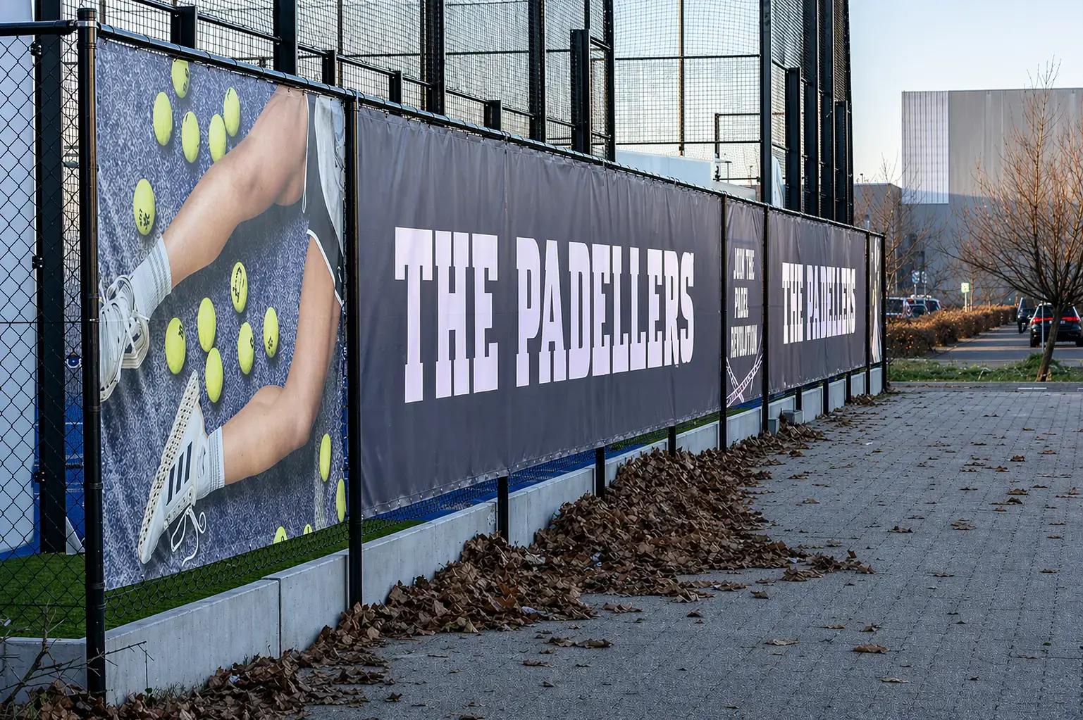 Lange hekwerkbanner met The Padellers-logo en padelbeeld langs een padelbaan, bevestigd aan zwart gaashekwerk als opvallende buitenreclame.