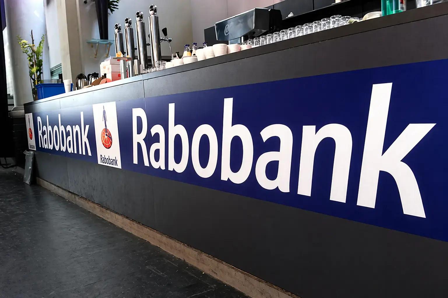 Blauwe Rabobank baliebelettering met witte logo’s en tekst op een donkere bar- of toonbankfront in een interieur.