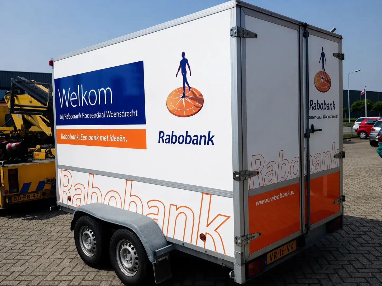 Dubbelassige gesloten aanhanger met full colour voertuigbelettering van Rabobank, inclusief welkomsttekst, logo en oranje-blauwe huisstijl op de zijkant en achterdeuren.