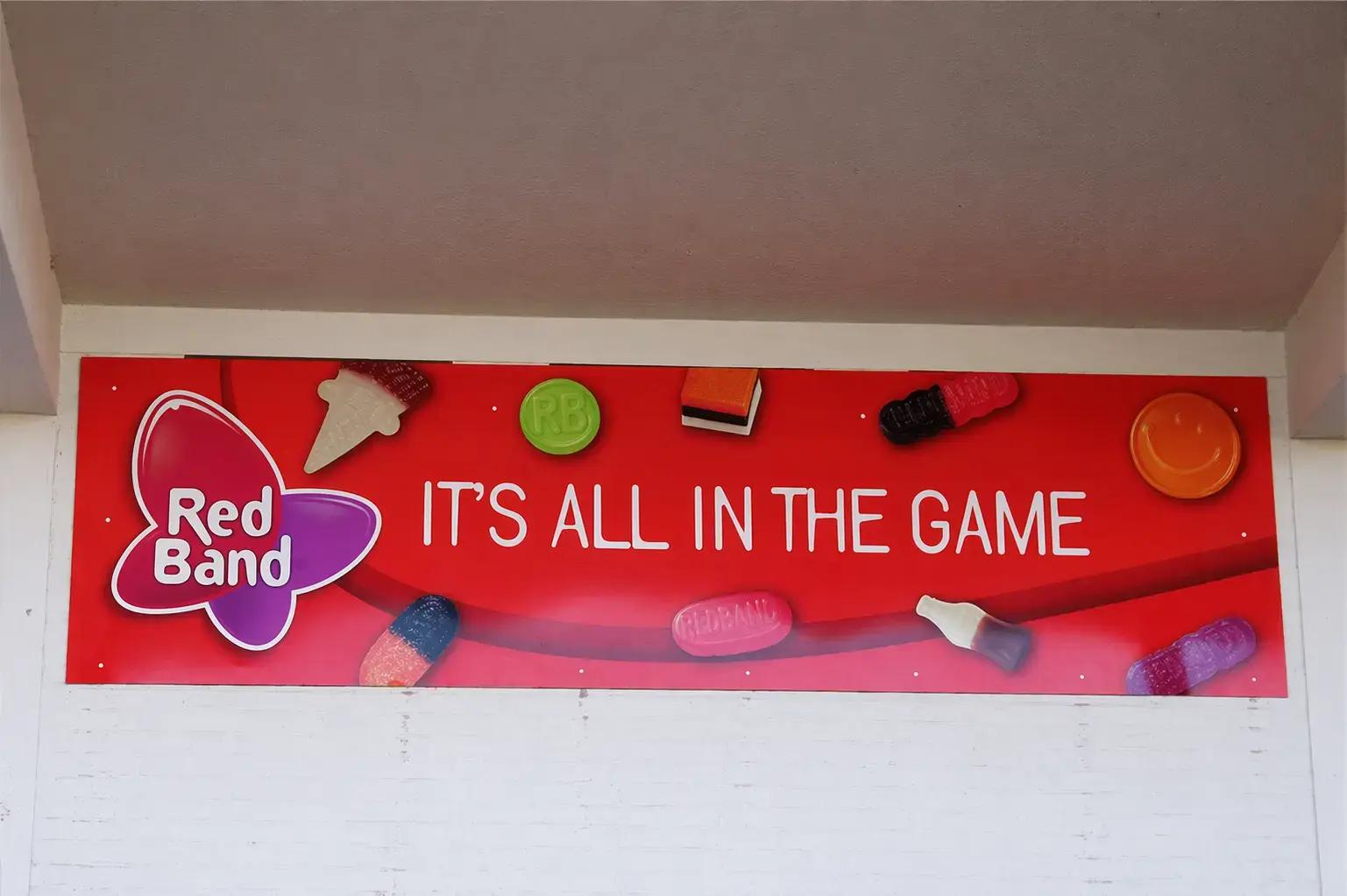 Rood reclamebord met Red Band logo, snoepafbeeldingen en de tekst IT’S ALL IN THE GAME boven een witte wand.