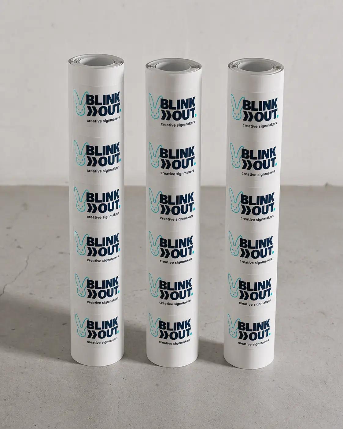Drie rollen witte etiketten met herhaald BLINK OUT creative signmakers-logo, gepresenteerd als bedrukte stickers op een neutrale ondergrond.