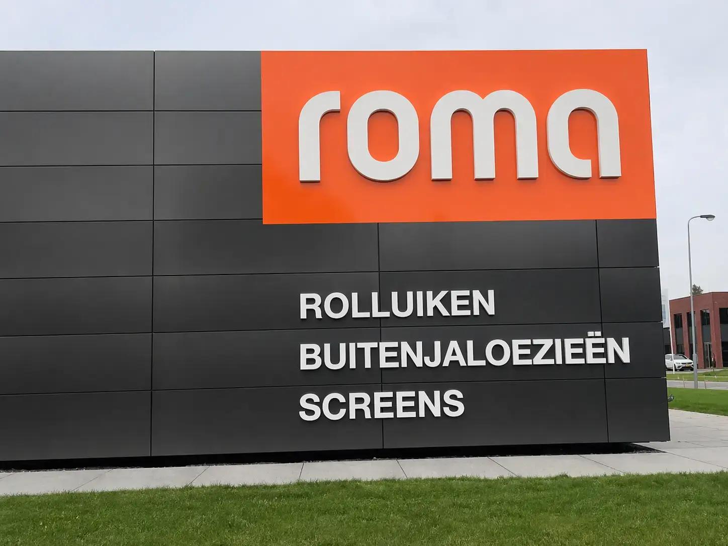 Moderne gevelreclame op zwarte gevelpanelen met oranje topvlak, witte 3D-letters van roma en productbelettering voor rolluiken, buitenjaloezieën en screens.
