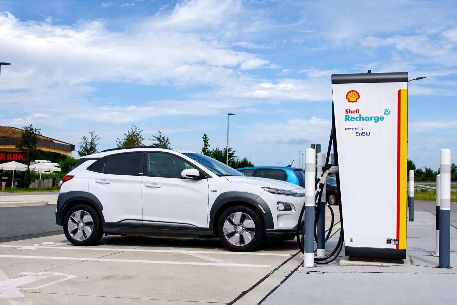 Shell Recharge laadpaal met branding bij een snellaadstation waar een witte elektrische auto wordt opgeladen.