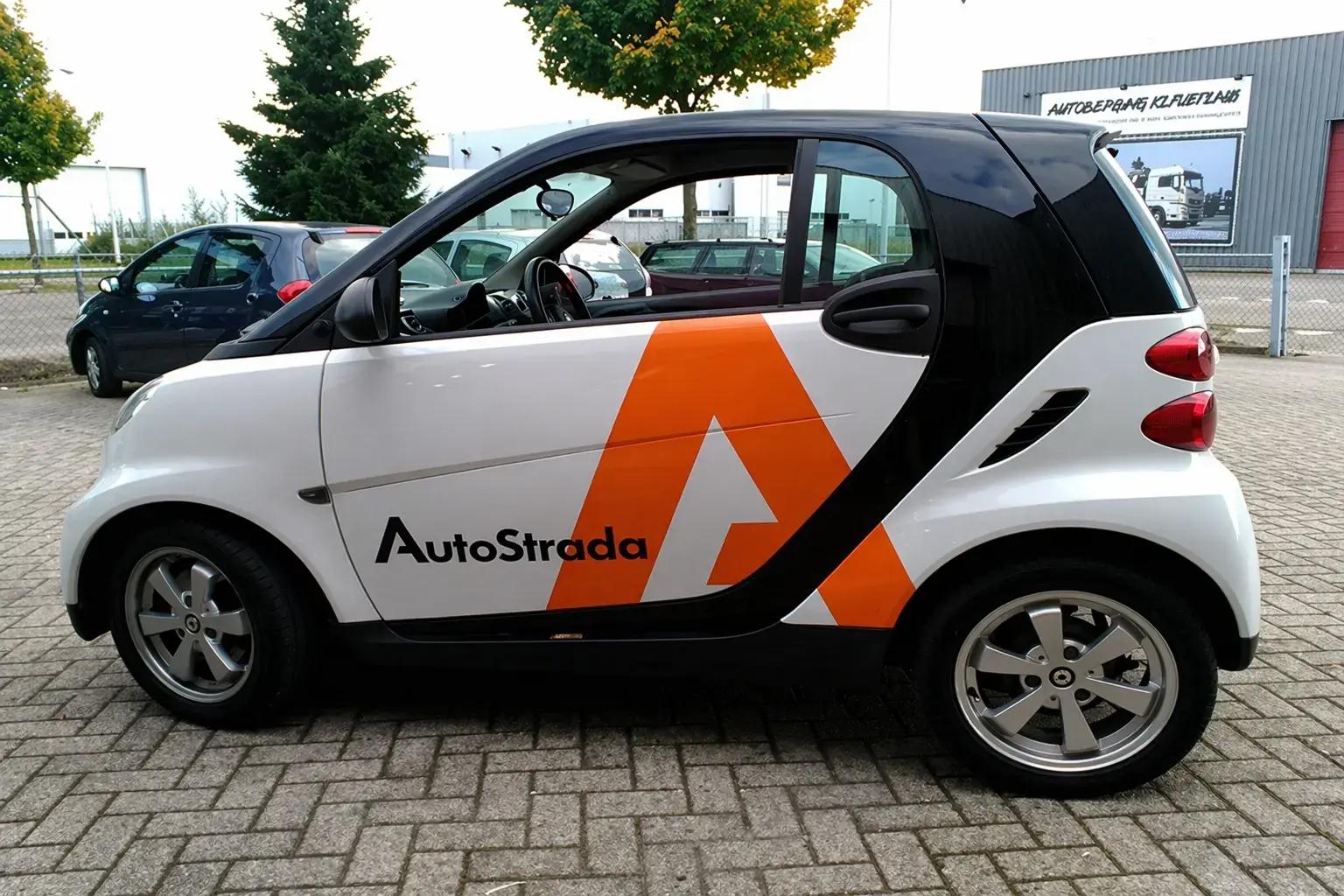 Zijaanzicht van een compacte witte stadsauto met zwarte panelen en opvallende oranje AutoStrada voertuigbelettering op de deur en achterzijde, geparkeerd op een bedrijventerrein.