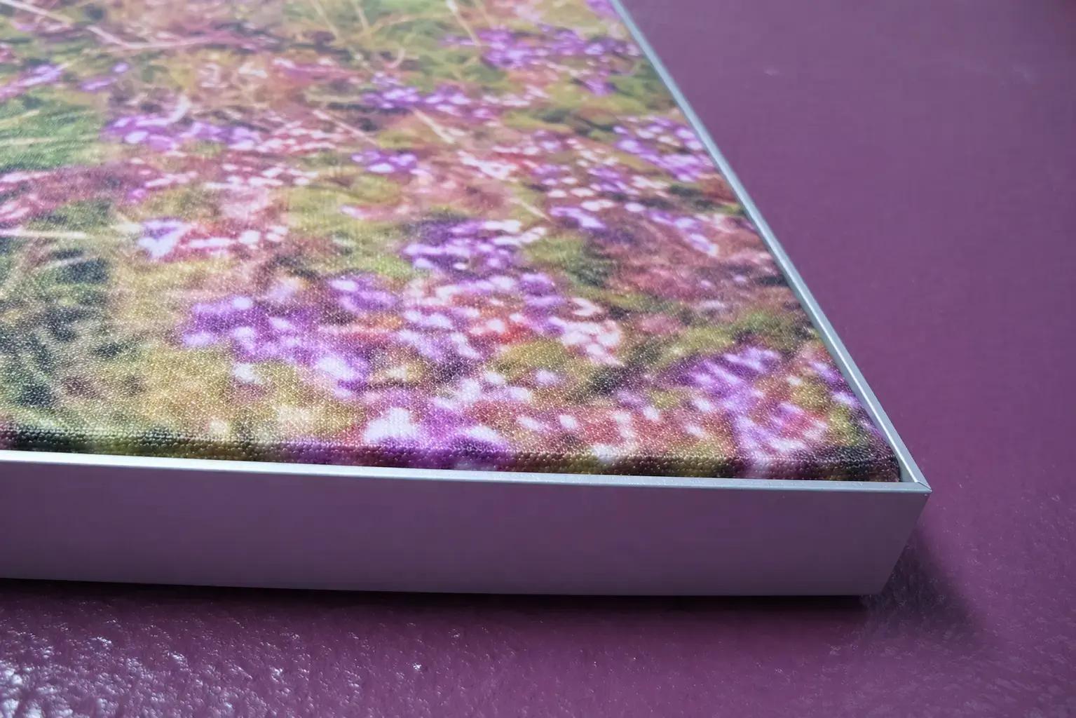 Close-up van een wanddecoratie met fotoprint van paarse bloemen in een strak aluminium frame, gemonteerd op een paarse structuurmuur.