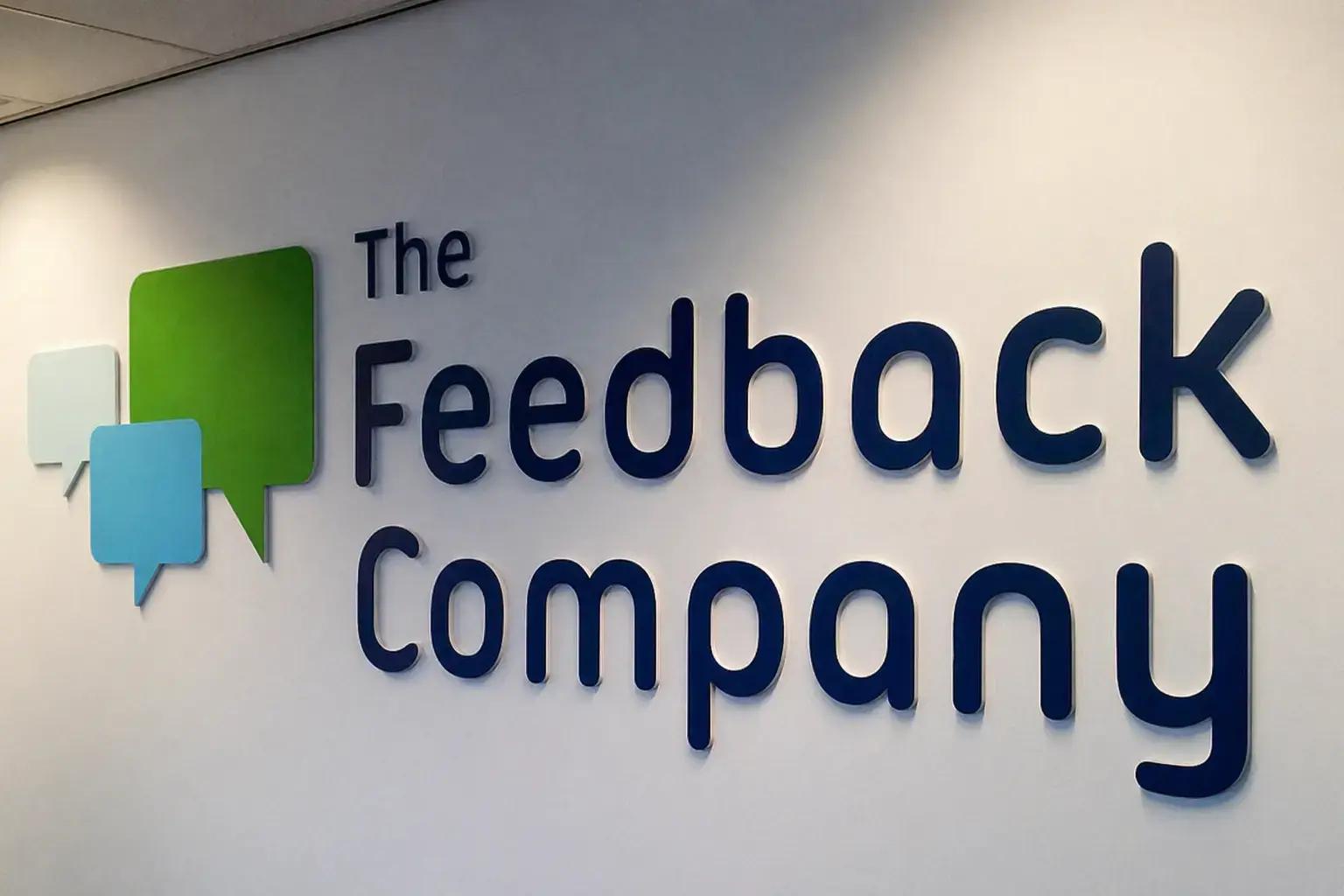 Wandlogo van The Feedback Company met donkerblauwe 3D letters en gekleurde tekstballon-iconen, gemonteerd op een witte binnenmuur als bedrijfsreclame.