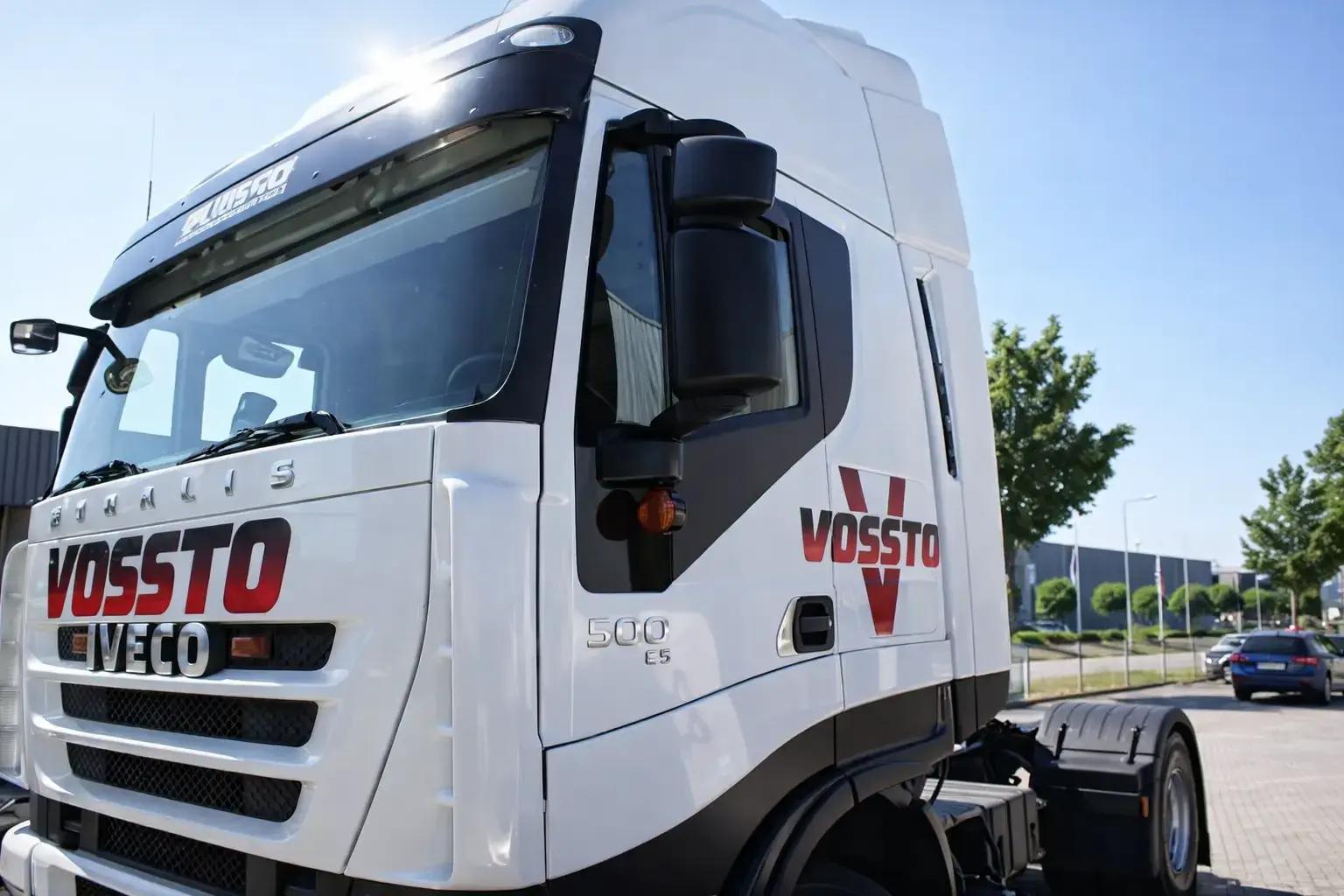 Witte vrachtwagencabine met VOSSTO- en IVECO-belettering in rood en zwart op front en portier, gefotografeerd op een bedrijventerrein.