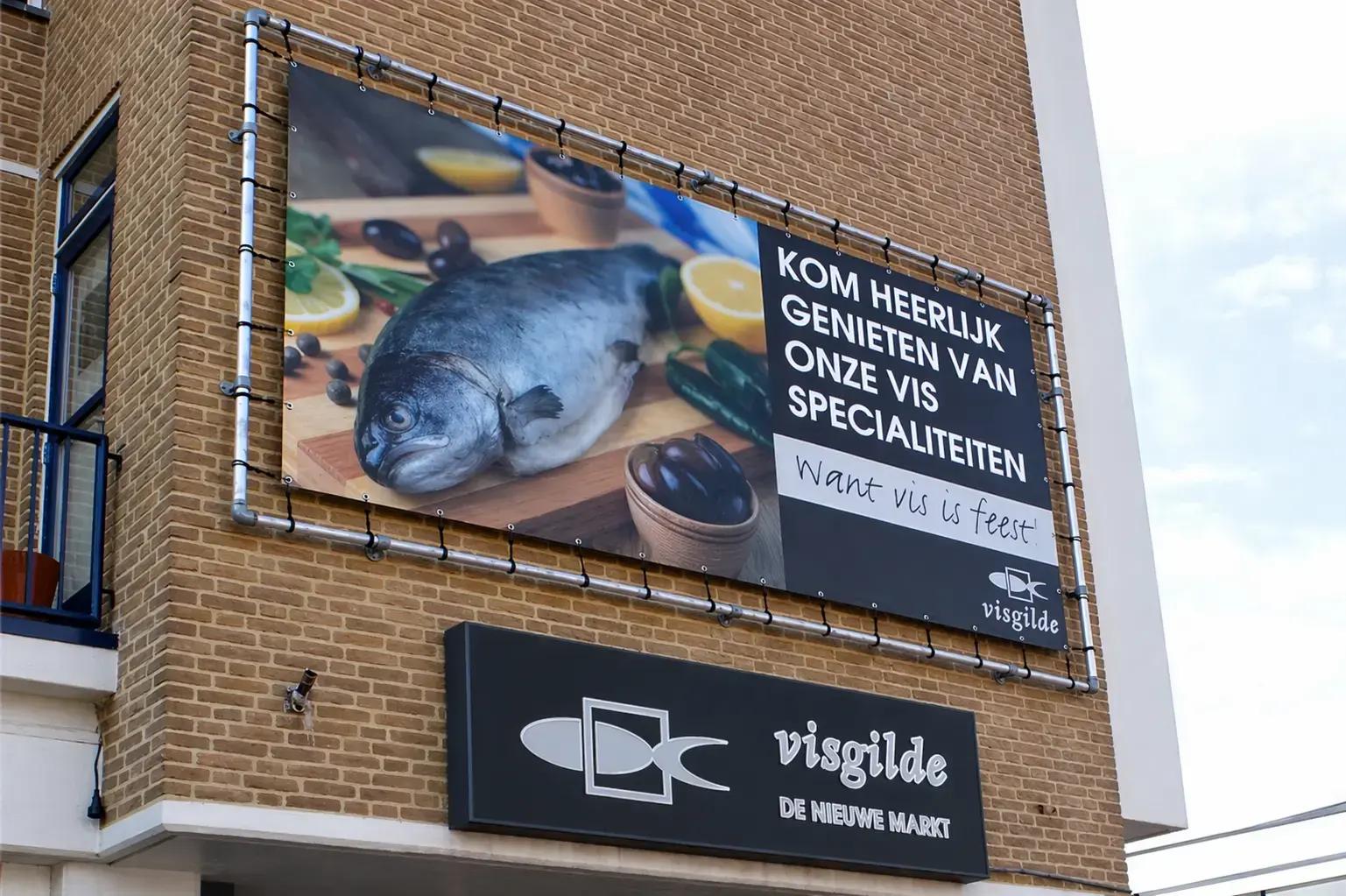 Gevelreclame met groot visbanner in buizenframe en zwart Visgilde bord op een bakstenen winkelgevel.
