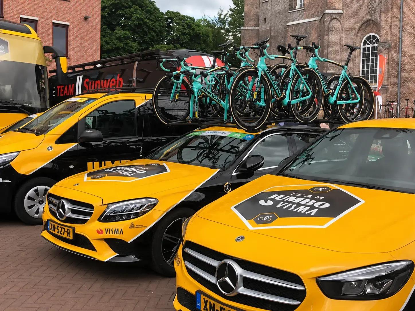 Gele en zwarte Team Jumbo-Visma teamwagens met opvallende autobelettering en dakreclame, voorzien van Bianchi racefietsen op dakdragers bij een bakstenen gebouw.