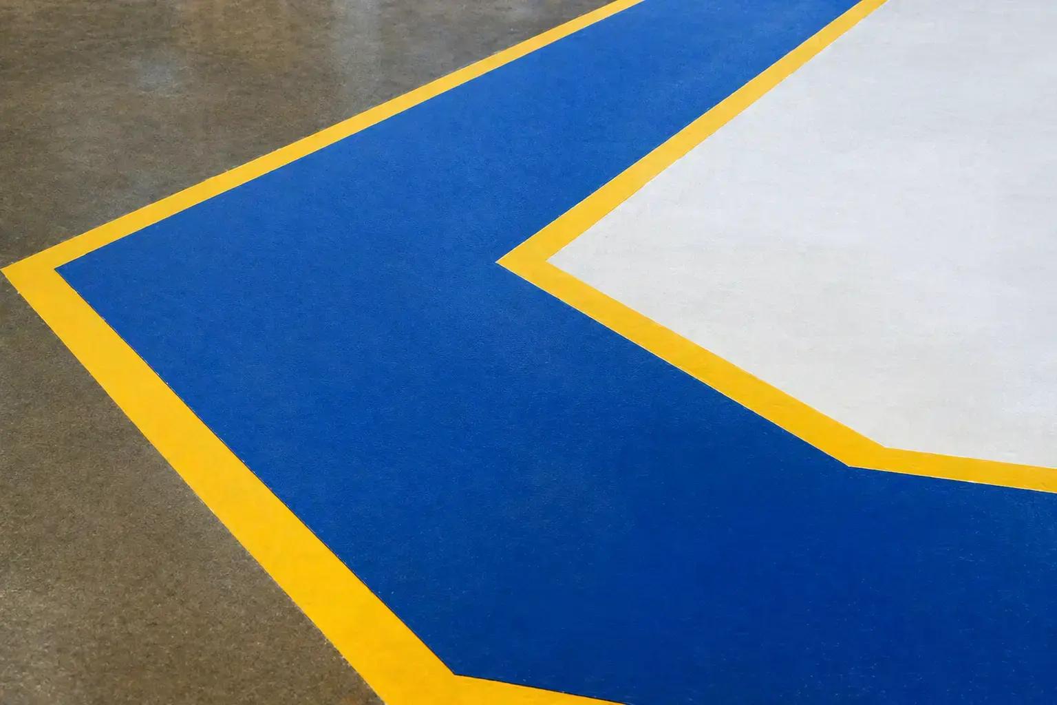 Blauwe vloerzone met gele belijning en witte markering op een industriële betonvloer, toegepast als duidelijke veiligheidsmarkering en routeaanduiding.