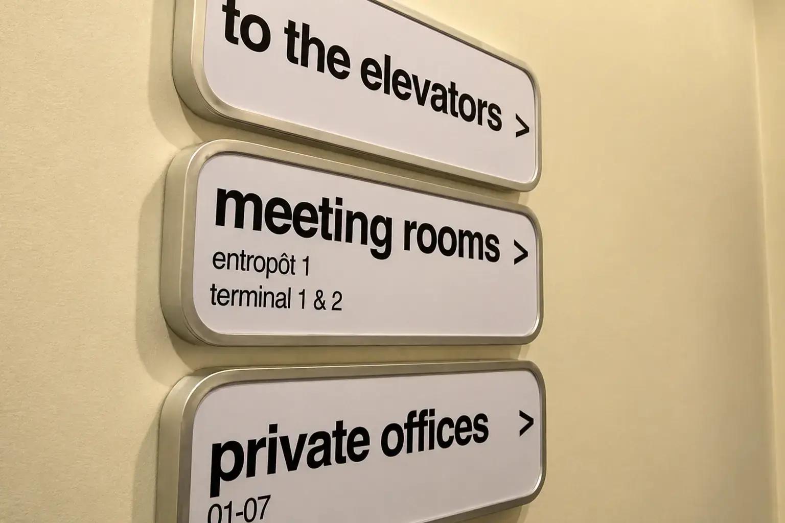 Wandgemonteerde wayfinding-borden met aluminium frame en zwarte tekst voor liften, vergaderruimtes en kantoren op een lichte binnenmuur.