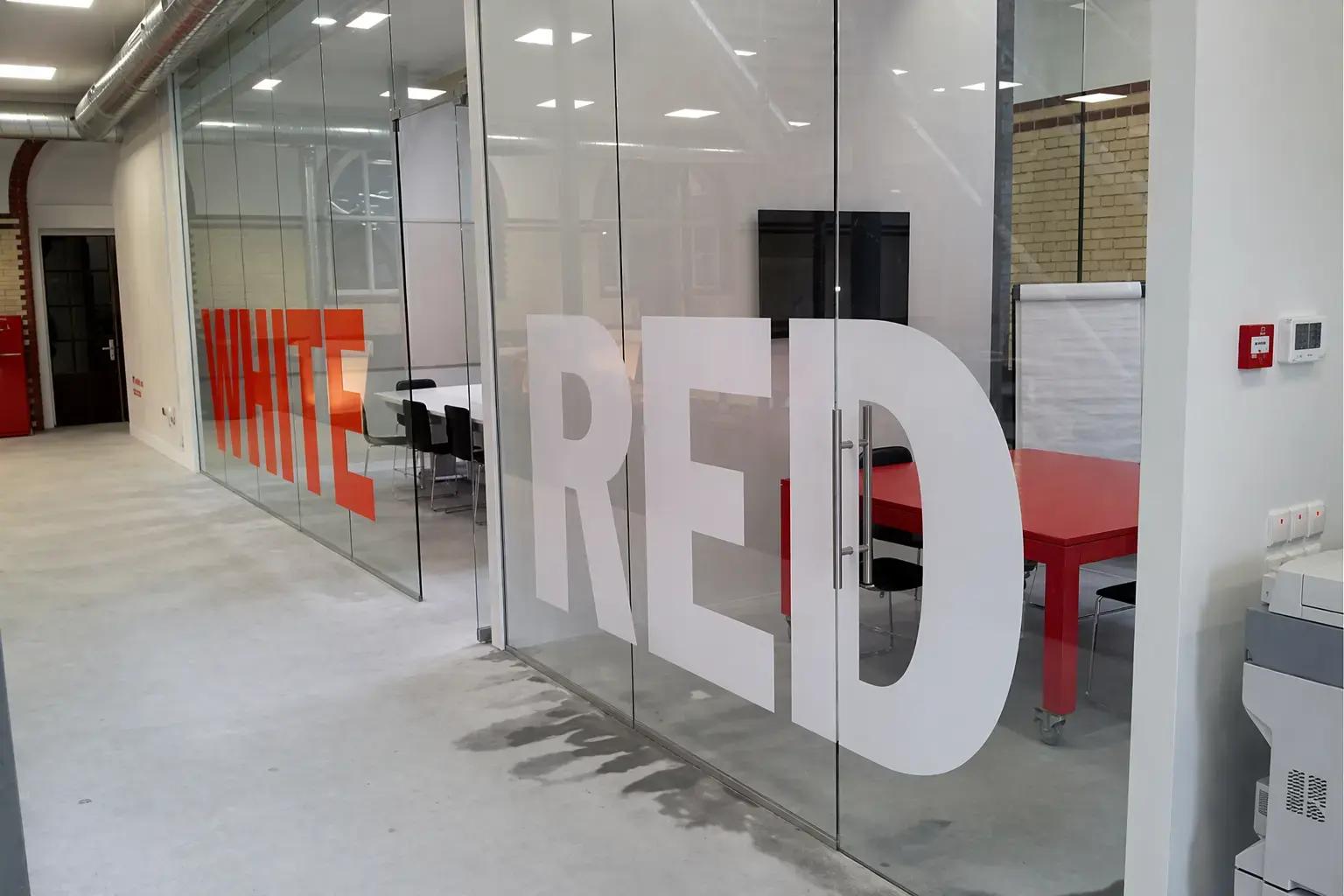 Glasfoliebelettering met grote witte en rode teksten op transparante kantoorwanden voor privacy en ruimte-aanduiding in een modern interieur.