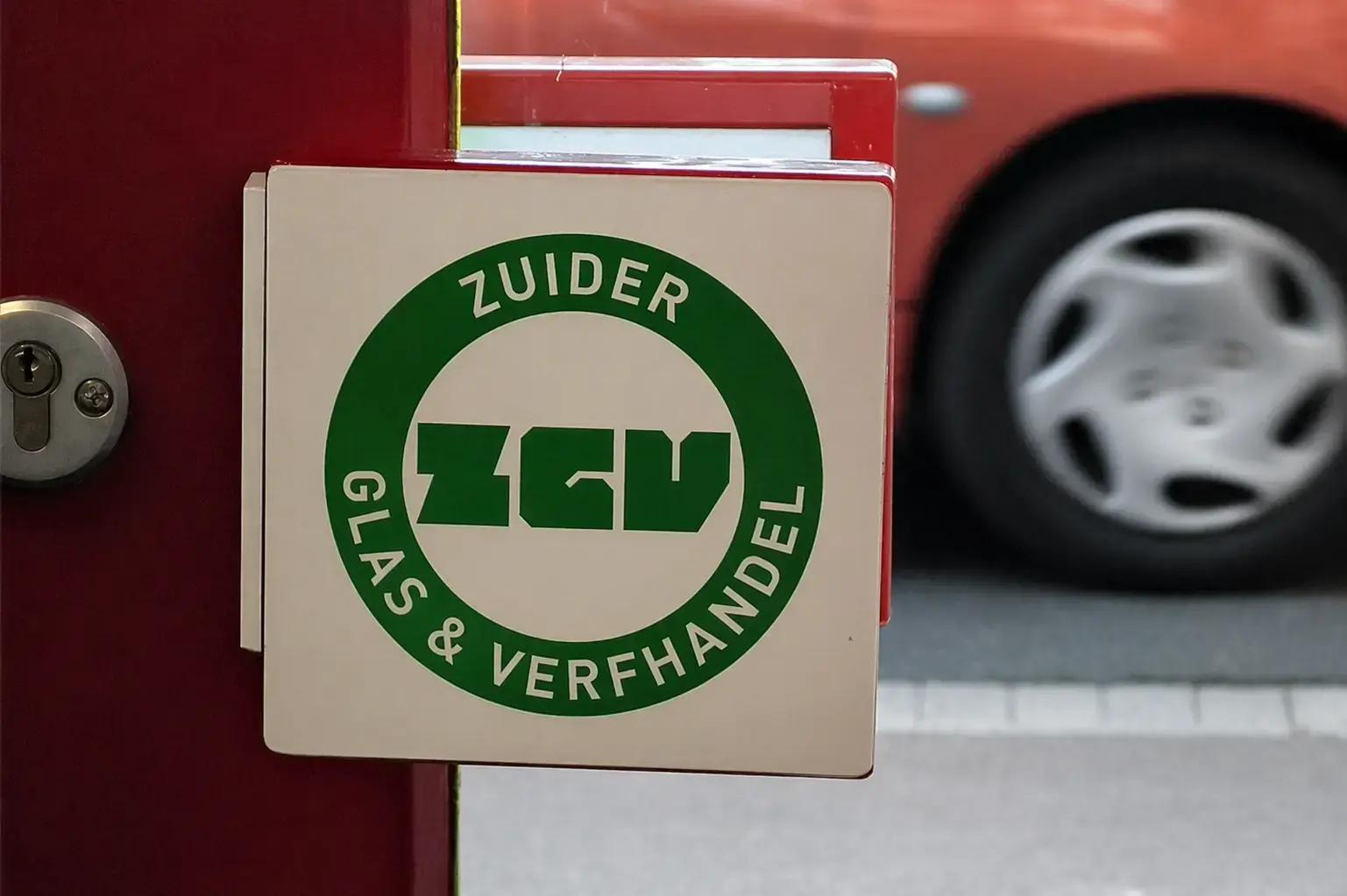 Uitstekend gevelbord met groen Zuider Glas & Verfhandel logo op een rood kozijn bij een winkelingang.