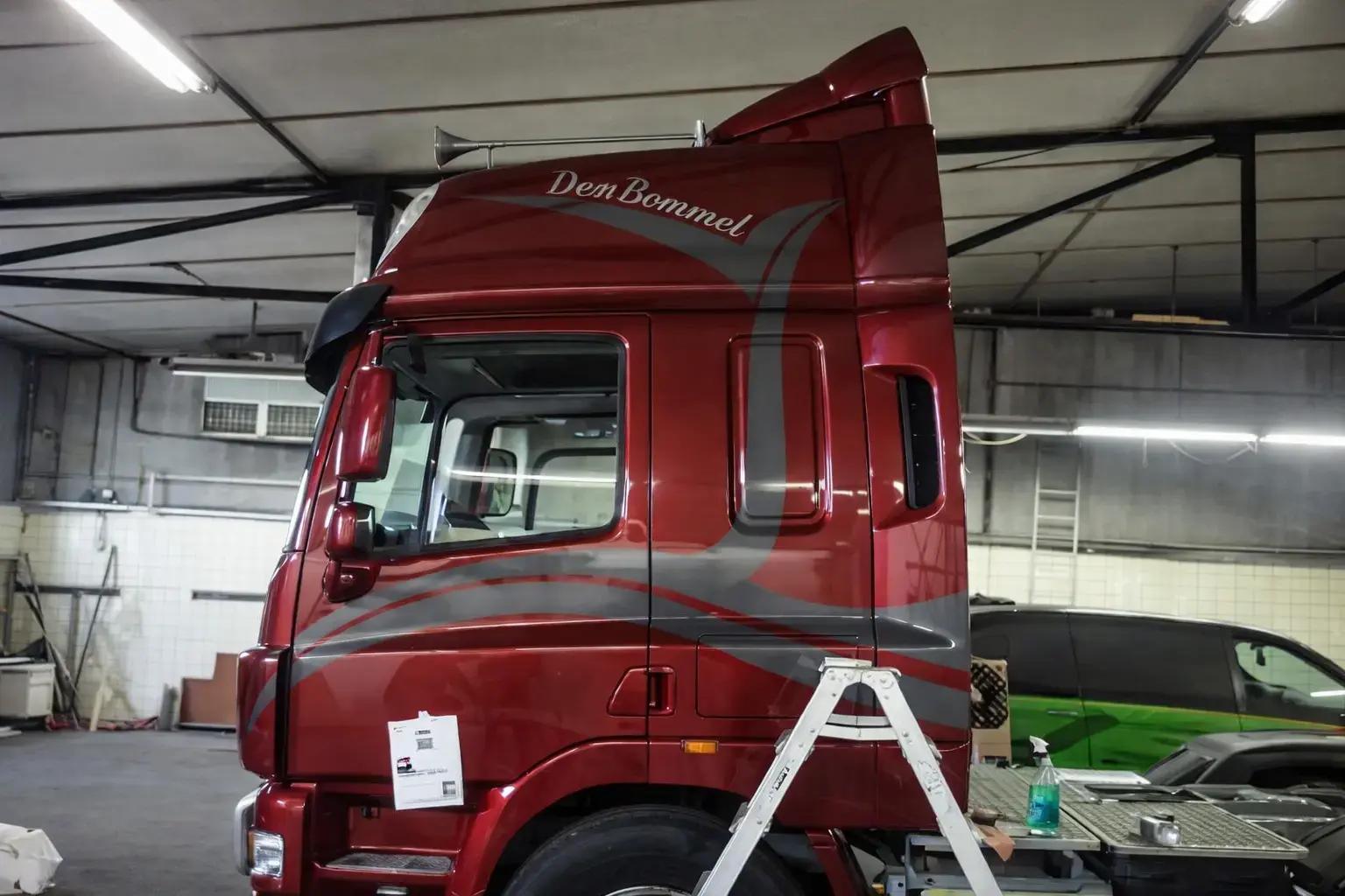 Bordeauxrode vrachtwagencabine met zilvergrijze striping en Den Bommel-belettering op de slaapcabine, gefotografeerd in een werkplaats.