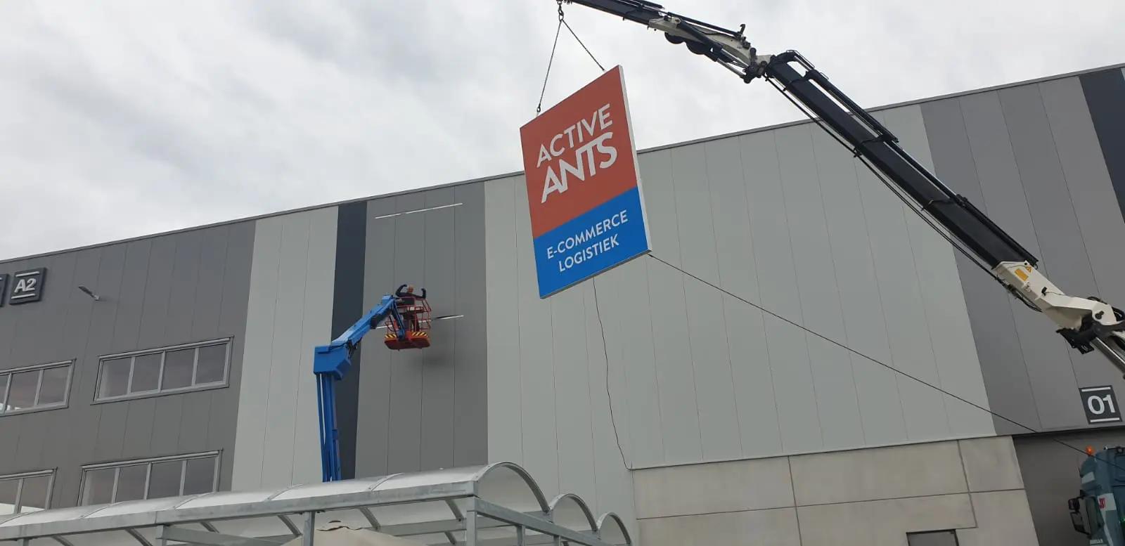 Kraan hijst de Active Ants-lichtbak naar de gevel, naast een blauwe hoogwerker.
