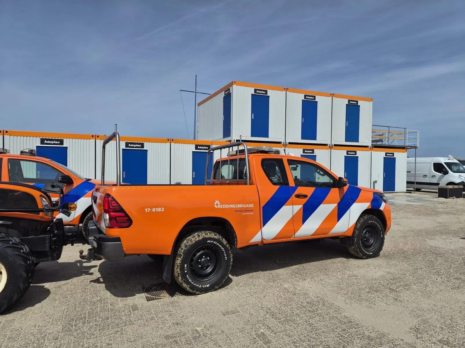 Strandpost met blauwe deuren, oranje dakrand en hulpdienstvoertuigen op de voorgrond.