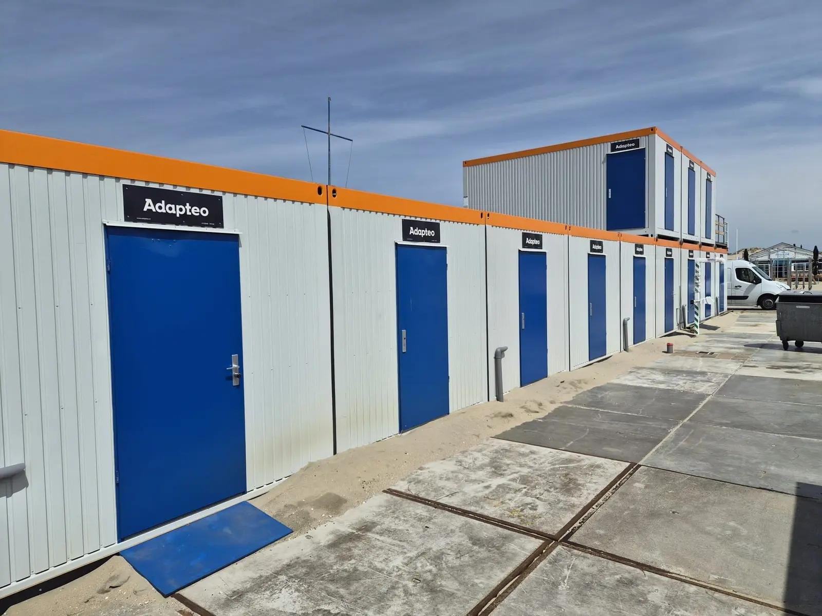 Blauwe deuren en oranje dakrand langs de modulaire strandpost.
