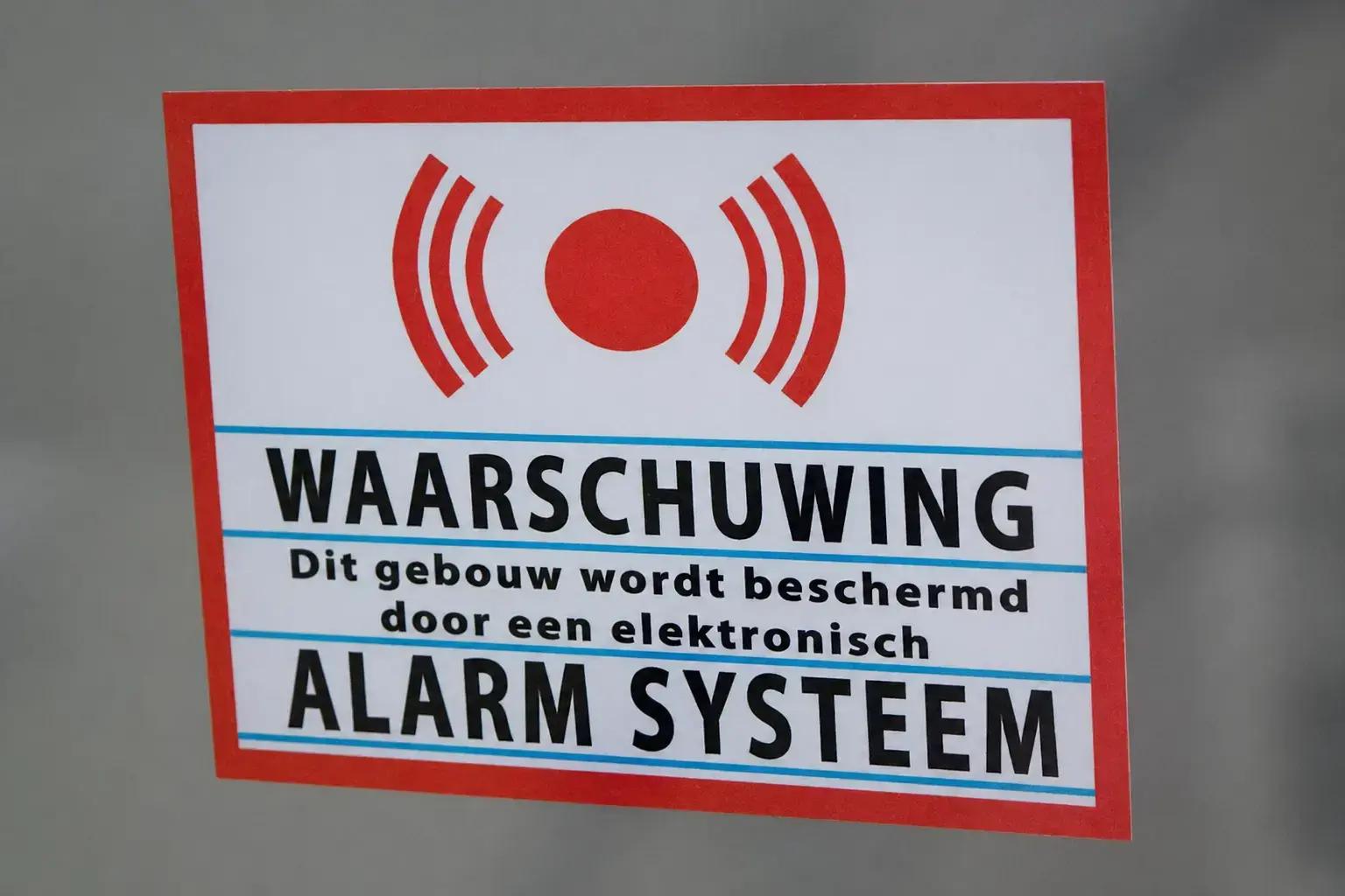 Waarschuwingssticker met rood alarmsymbool en tekst over elektronisch alarmsysteem, aangebracht op een grijze ondergrond als beveiligingssignalering.