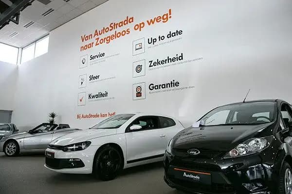 Interieur van een autoshowroom met grote wandbelettering en informatieve muurgrafiek van AutoStrada, met kernwaarden in zwarte en oranje tekst boven meerdere showroomauto’s.