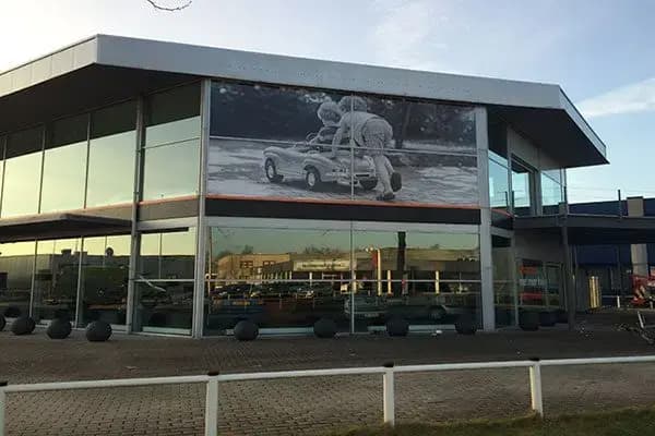 Moderne glazen showroomgevel met grote zwart-wit gevelbanner over de bovenpui en subtiele horizontale accentstrip, geplaatst aan een hoekpand op een bedrijventerrein.