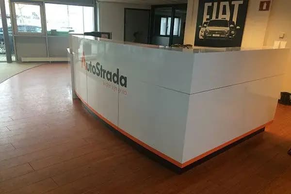 Witte ontvangstbalie met AutoStrada-logo in de showroom, uitgevoerd als strakke interieurbelettering met subtiele oranje accentlijn op de voorzijde.