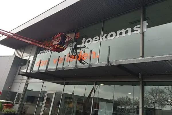 Grote witte en oranje raambelettering op de glazen gevel van een bedrijfspand, gemonteerd met een hoogwerker aan de bovenverdieping.