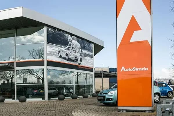Oranje vrijstaande reclamemast met AutoStrada-logo voor een autobedrijf, naast een glazen showroomgevel met grote raambestickering en gevelbanner.