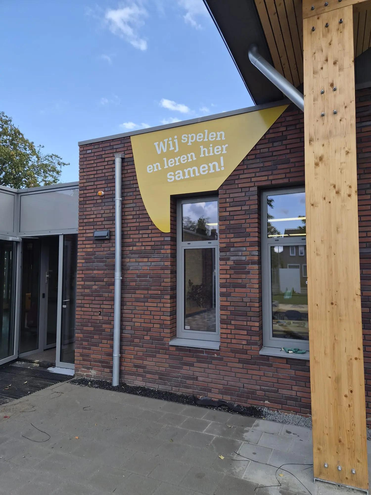Geel gevelpaneel met de tekst Wij spelen en leren hier samen bij De Baayaert in Wouw.