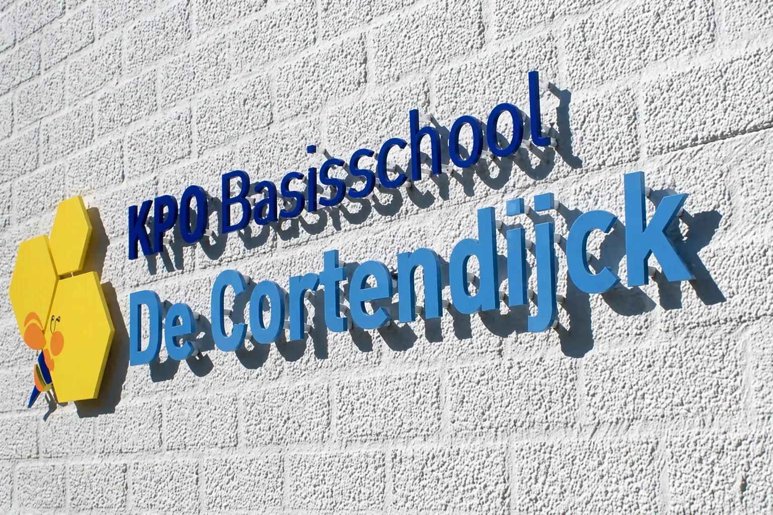 Witte gevel met blauwe 3D-belettering van KPO Basisschool De Cortendijck en een geel zeshoekig logo met bij, gemonteerd als opvallende schoolsigning.