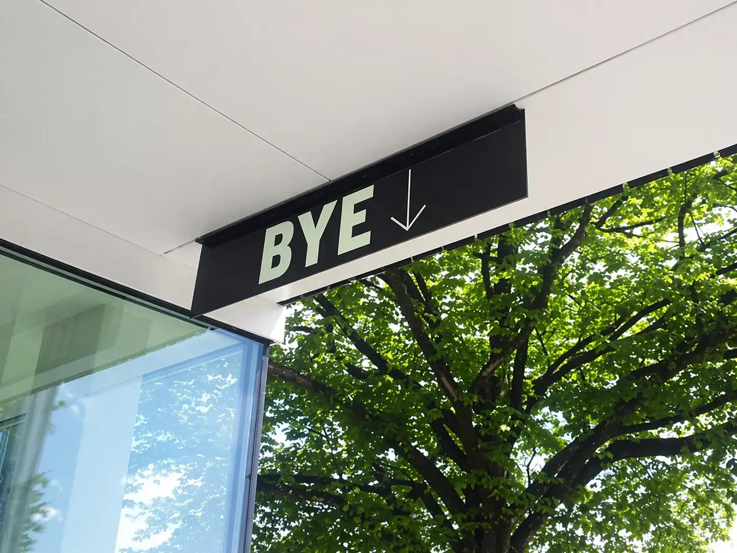 Zwart hangend wayfinding-bord met de tekst BYE en een pijl omlaag, gemonteerd onder een luifel bij een glazen gevel.