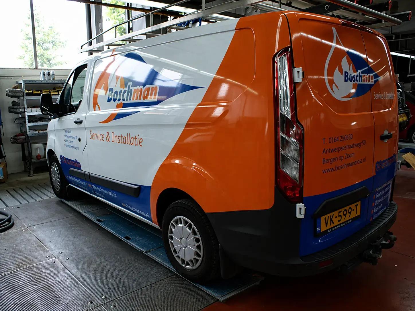 Bestickerde bedrijfsbus met oranje-wit-blauwe voertuigbelettering, Boschman-logo en de tekst Service & Installatie, gefotografeerd in een werkplaats.