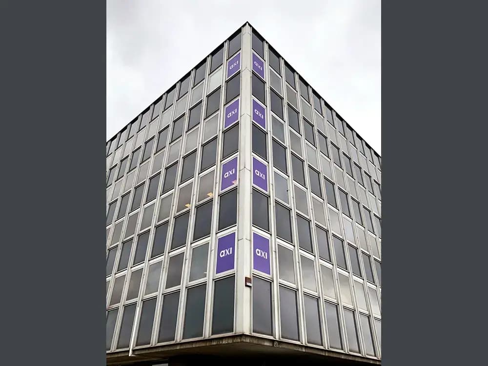 Hoekgevel van een modern kantoorgebouw met paarse raamstickers met axi-logo, verticaal aangebracht over meerdere verdiepingen als gevel- en raambelettering.