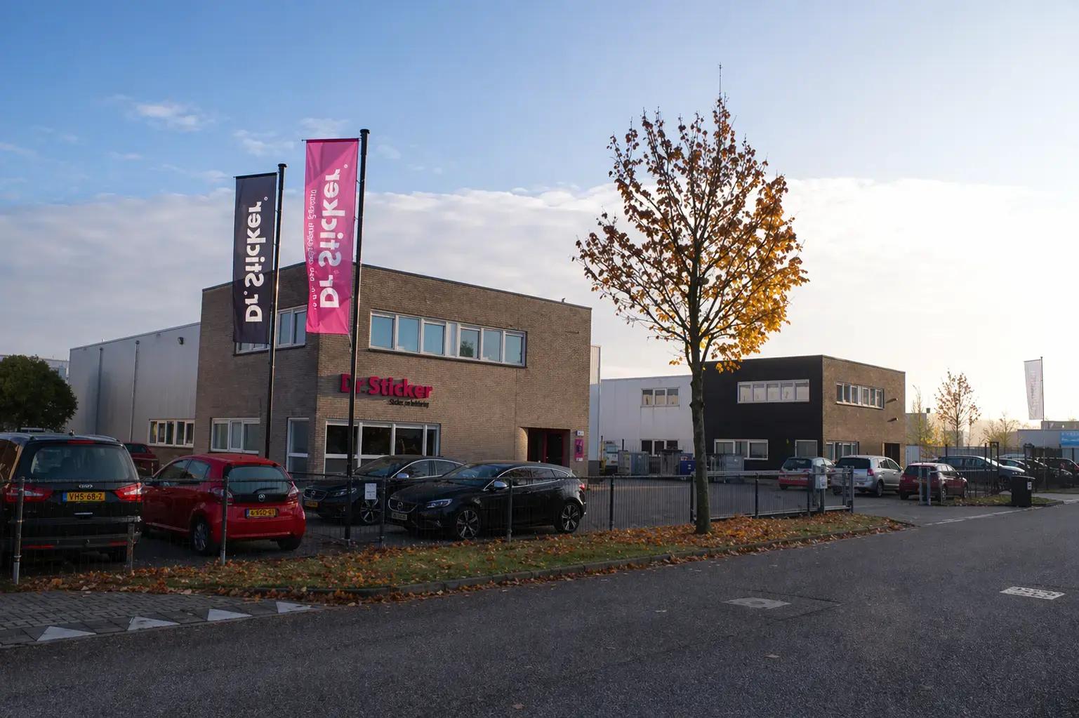 Bedrijfspand op een bedrijventerrein met gevelreclame van Dr. Sticker en twee hoge mastvlaggen in donkerblauw en magenta, gefotografeerd langs een straat met geparkeerde auto’s.