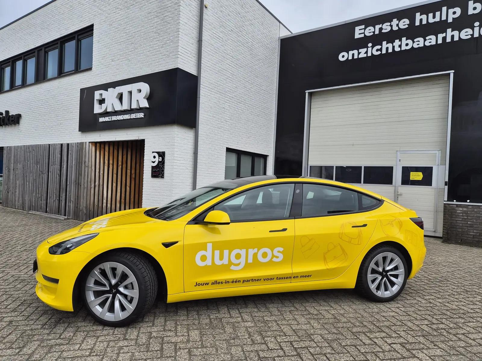 Zijaanzicht van de gele Dugros Tesla met groot wit logo en subtiele tasillustraties op de achterste carrosseriedelen.