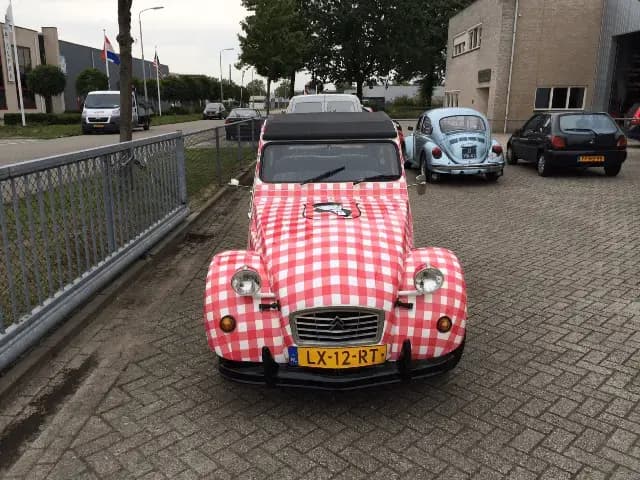Vooraanzicht van een klassieke auto met volledige voertuigwrap in rood-wit ruitpatroon, zwart cabriodak en illustratielogo op de motorkap, geparkeerd op een bedrijventerrein.