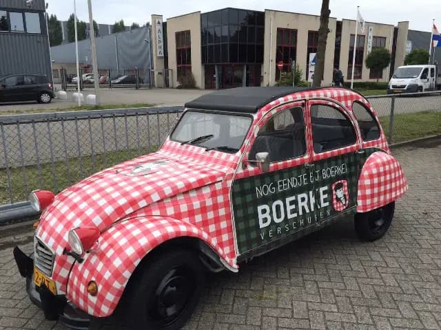 Klassieke auto met volledige voertuigwrap in rood-wit ruitpatroon en zwarte cabriokap, voorzien van groene zijbelettering met BOERKE-logo en promotionele tekst op een bedrijventerrein.