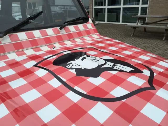 Close-up van de motorkap van een klassieke auto met rood-wit geruite voertuigwrap en groot zwart-wit illustratielogo op de voorzijde.