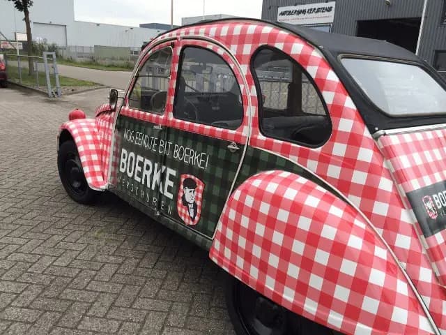 Klassieke auto met opvallende voertuigwrap in rood-wit ruitpatroon en zwarte cabriokap, voorzien van groene zijbelettering met BOERKE-logo en promotionele tekst op een bedrijventerrein.