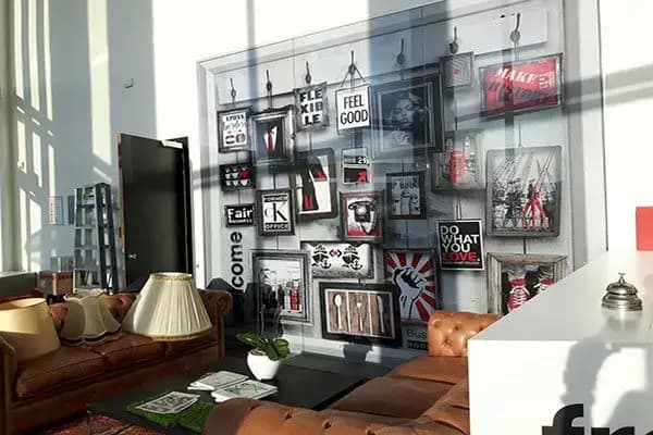 Loungehoek in een modern bedrijfsinterieur met leren banken en een grote full-color glaswandprint in collage-stijl met typografie, posters en framed artwork.