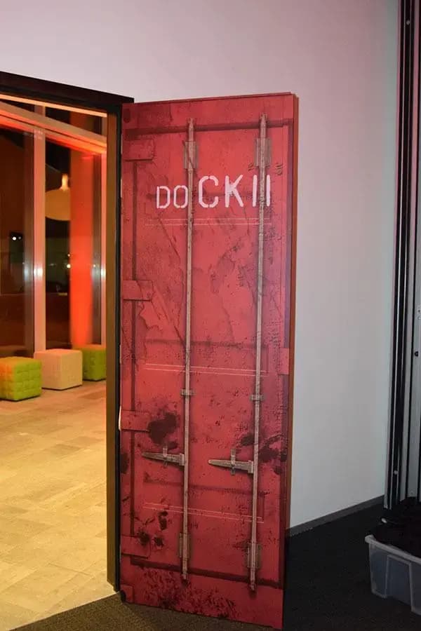 Rode deurprint in containerstijl met witte typografie naast een doorgang in een modern interieur met industriële uitstraling.