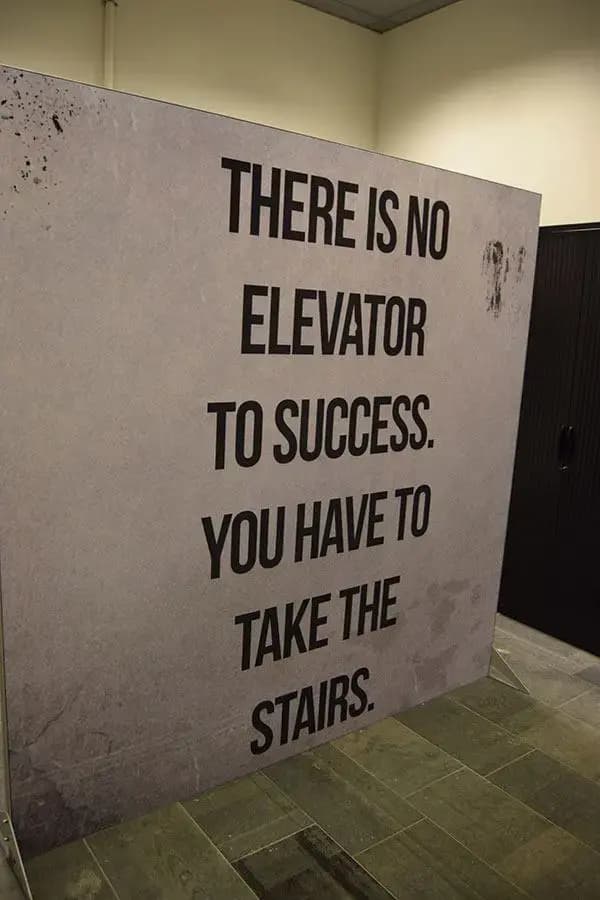 Vrijstaand grootformaat tekstpaneel met motiverende typografie “There is no elevator to success, you have to take the stairs” in een modern kantoorinterieur.