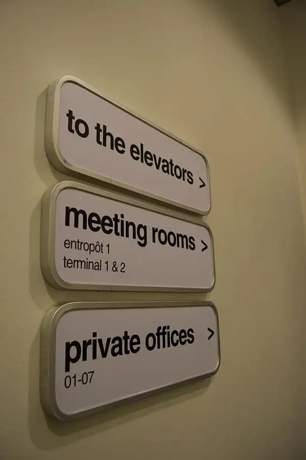 Witte wayfinding-bewegwijzering met afgeronde panelen voor liften, vergaderruimtes en private offices, gemonteerd op een lichte binnenwand.