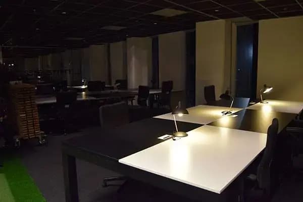 Donker kantoorinterieur met moderne werkplekken, bureaulampen op grote tafels en stoelen in een strak ingerichte open kantoorruimte.