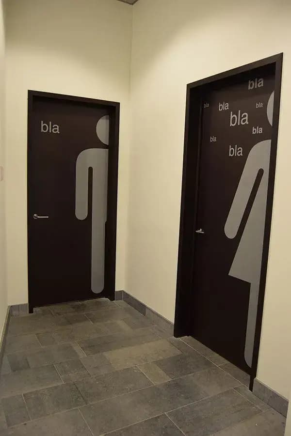 Twee donkere binnendeuren met grote grijze deurfolie in figuurvorm en typografische belettering, geplaatst als decoratieve wayfinding in een modern kantoorinterieur.