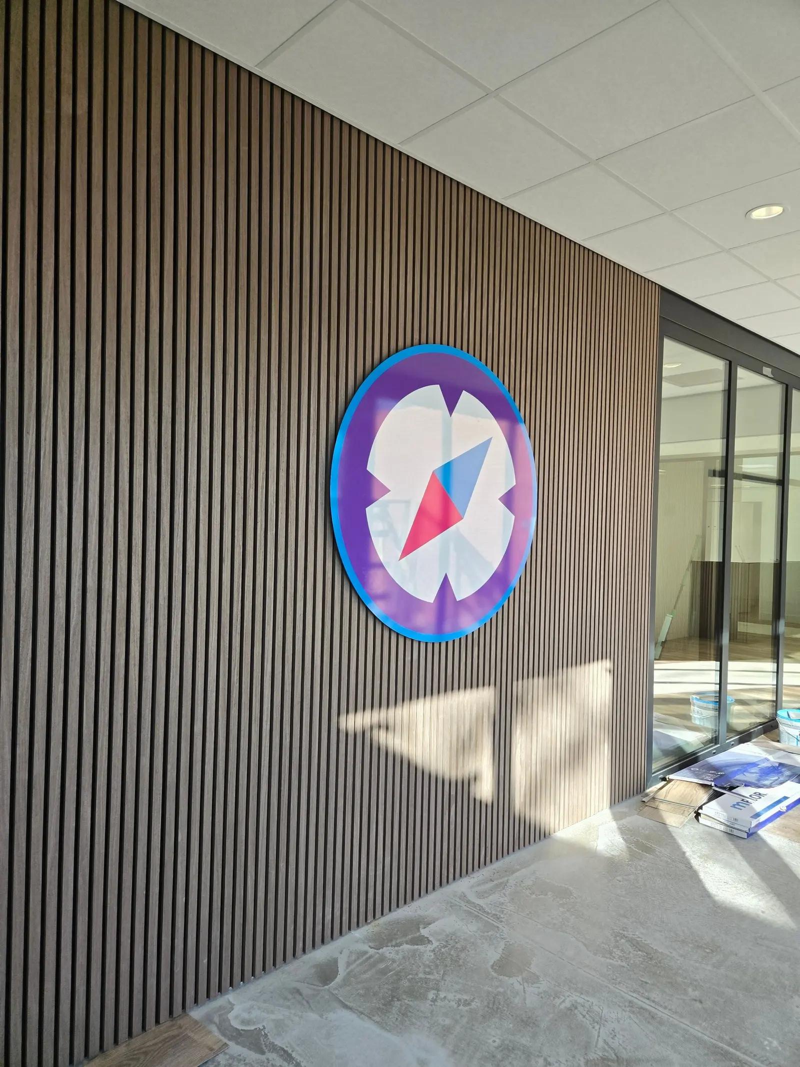 Ruimtelijk kompaslogo van FysioFaster op een houten lattenwand in het interieur.