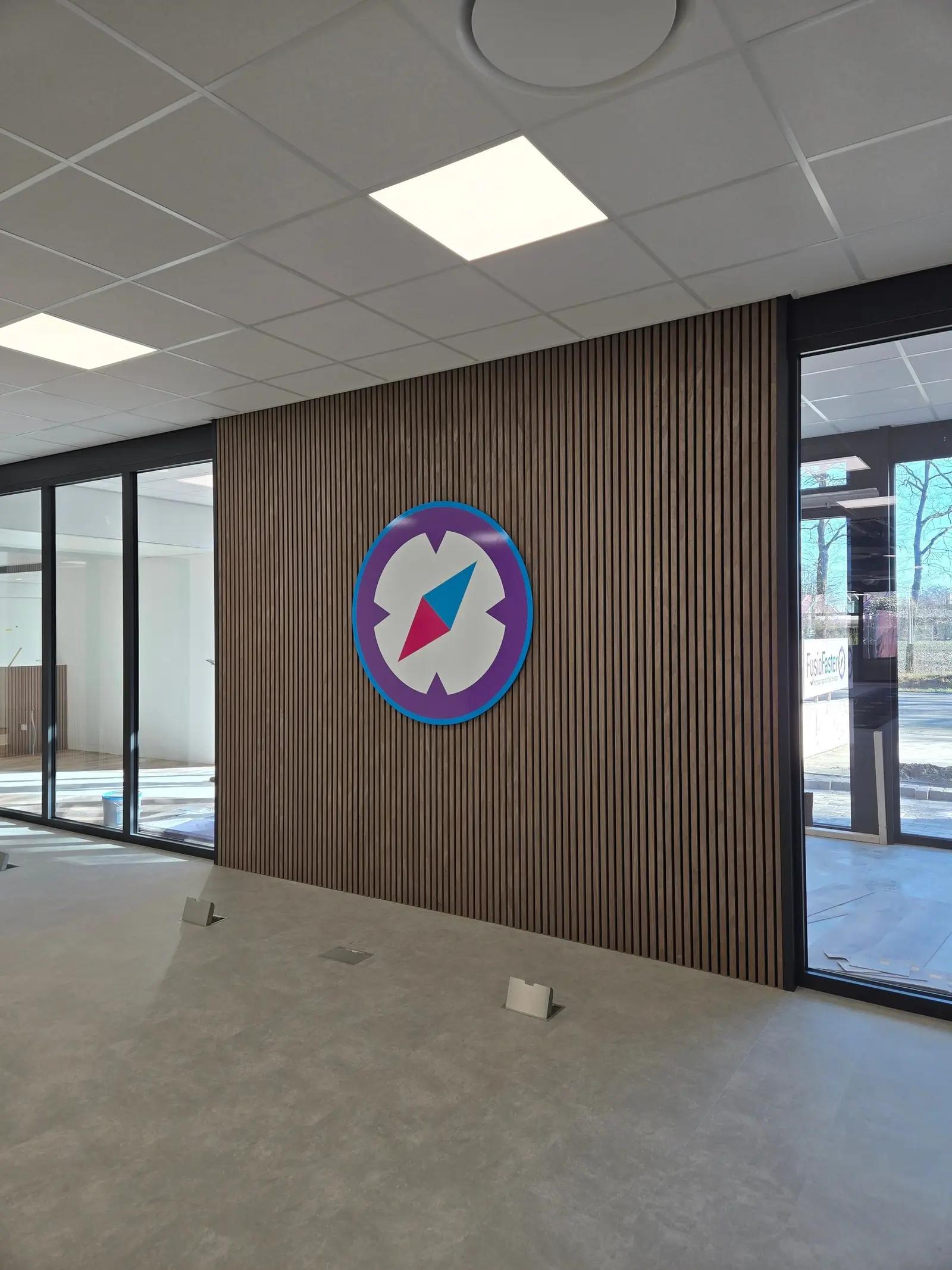 Kompaslogo op de lattenwand tussen glazen wanden in de FysioFaster-praktijk.